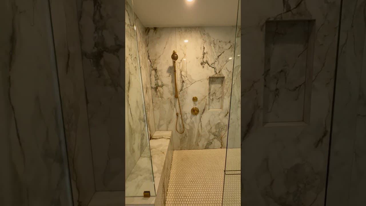 Marble clad shower behind frameless glass with brushed gold fittings, a stone bench and a penny round floor