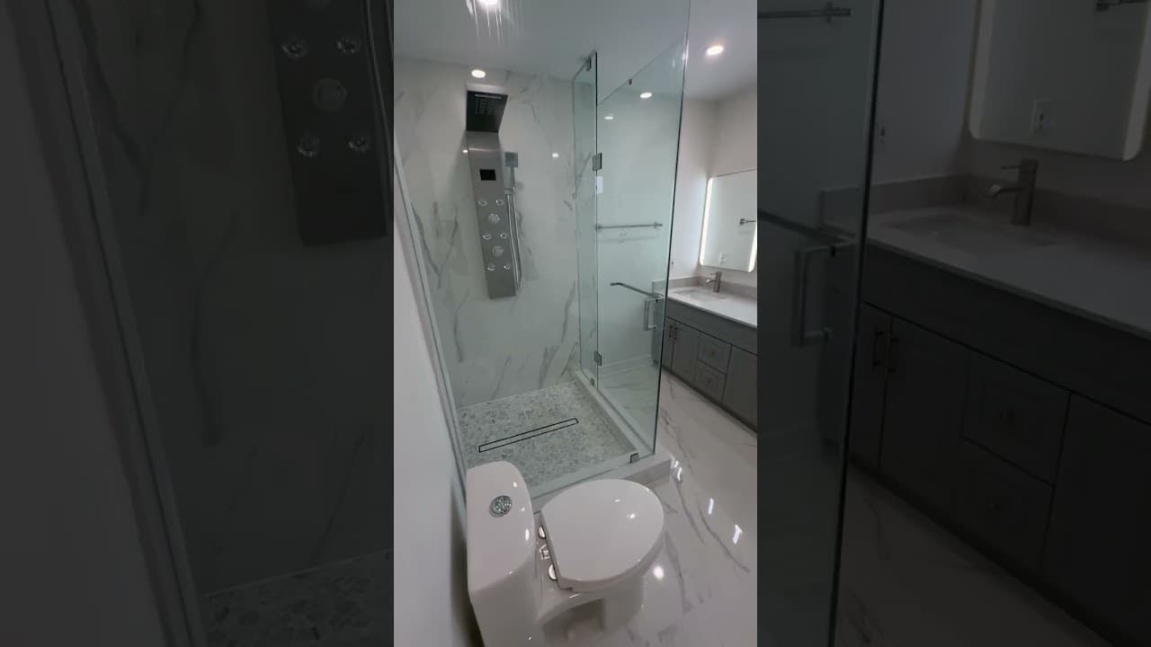 Frameless glass shower with a stainless shower panel and pebble floor, beside a dark vanity with a backlit mirror