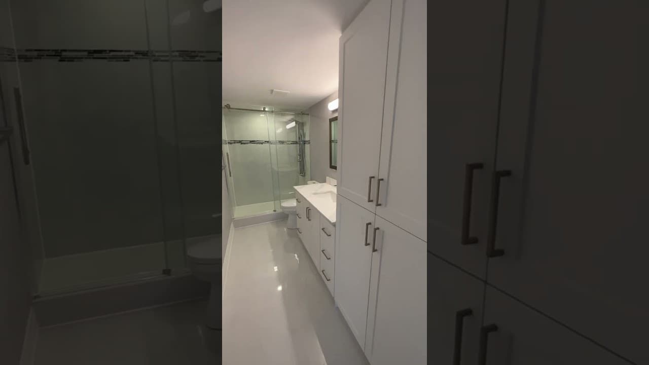 Narrow bathroom with a sliding glass shower, dark mosaic accent band, white vanity and a tall linen tower