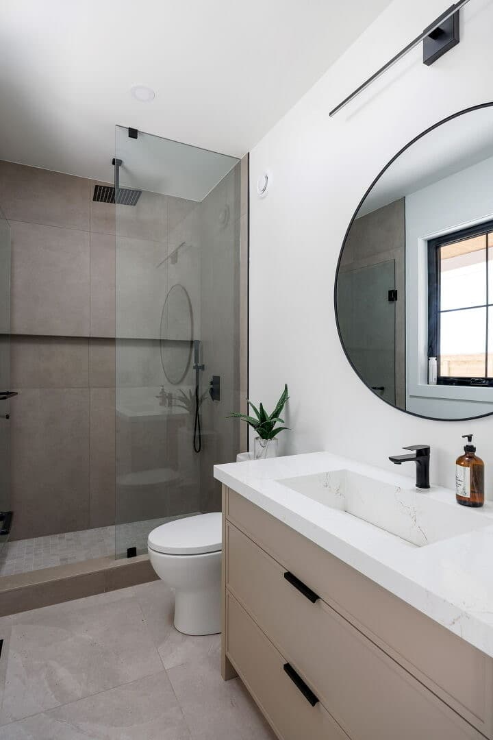 Walk in shower in concrete look tile with black fixtures, beside a round black mirror and taupe vanity