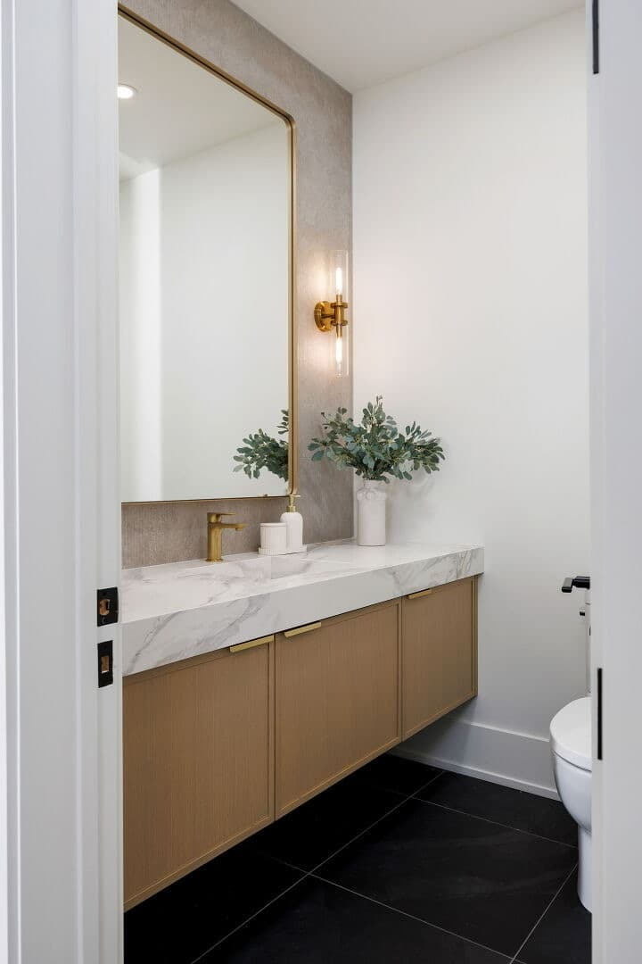 Powder room with a floating oak vanity, marble waterfall top, brass mirror and sconce over black floor tile