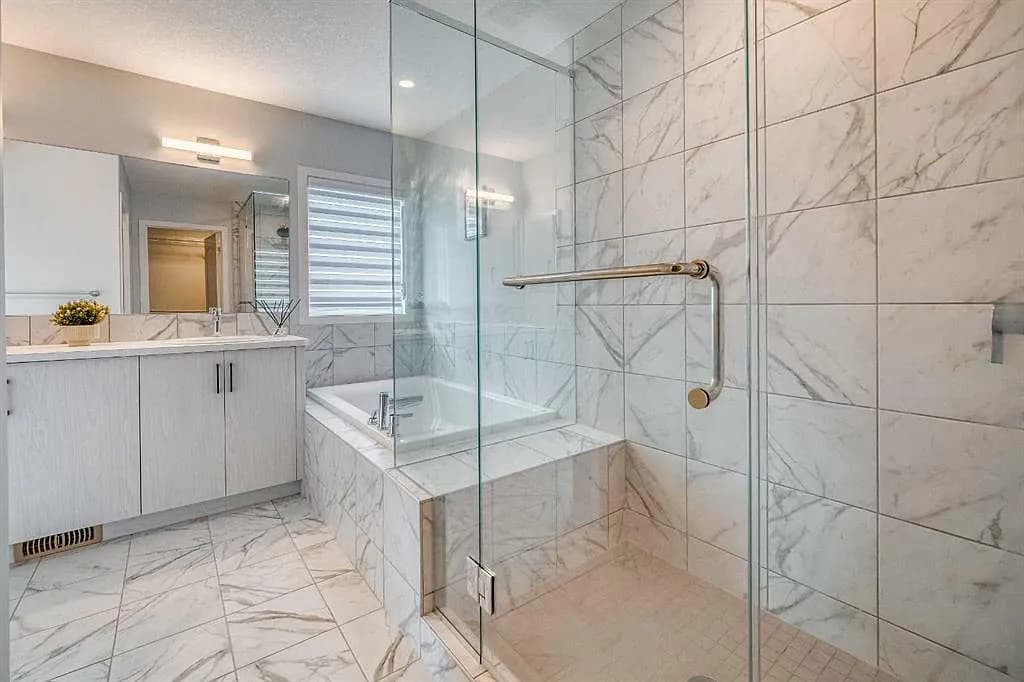 Marble tiled bathroom with a frameless glass walk in shower, a built in tub on a tiled surround, and a light wood vanity with a full width mirror