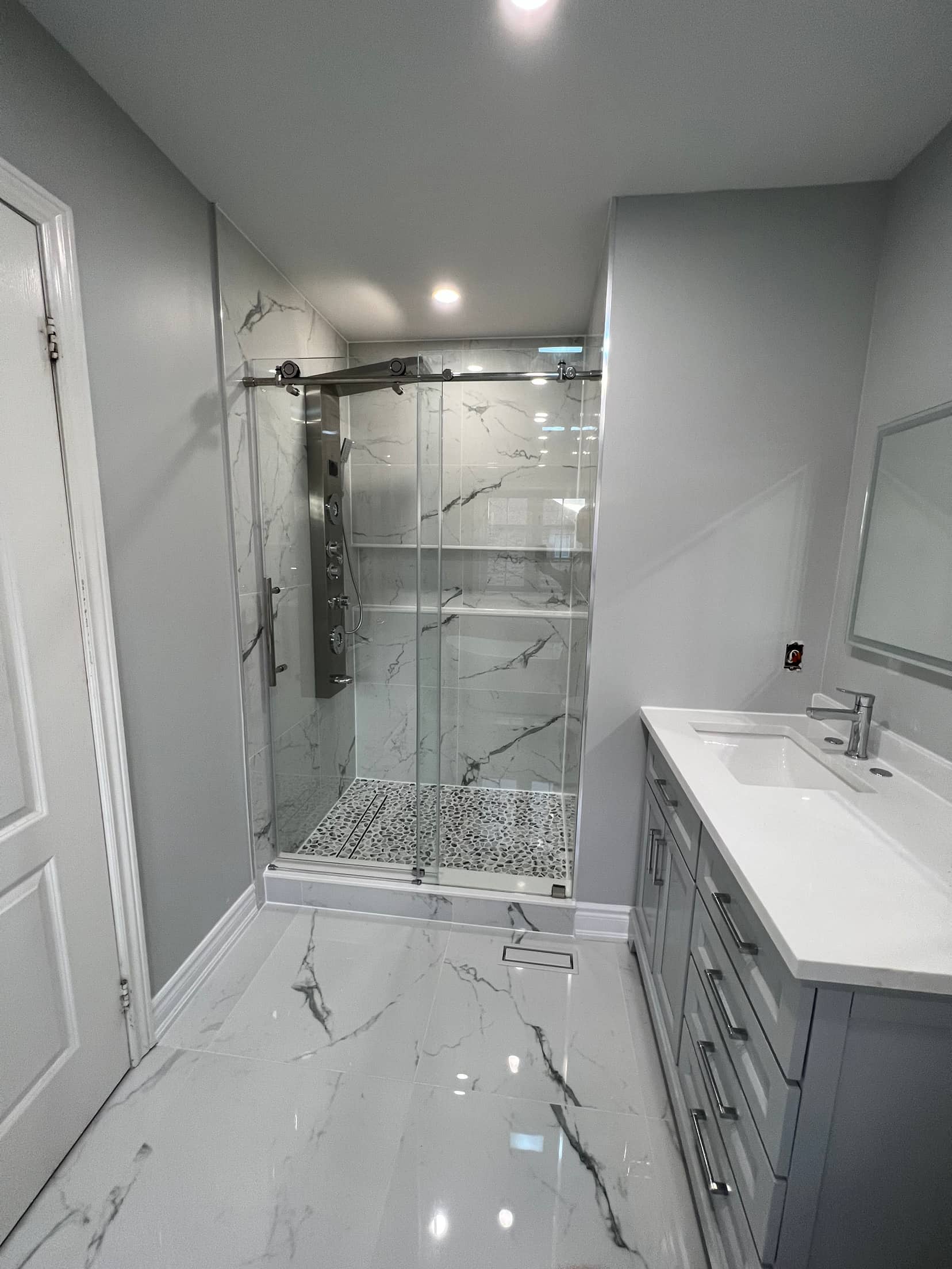 Marble tiled shower with a stainless shower panel and pebble mosaic floor, beside a grey vanity