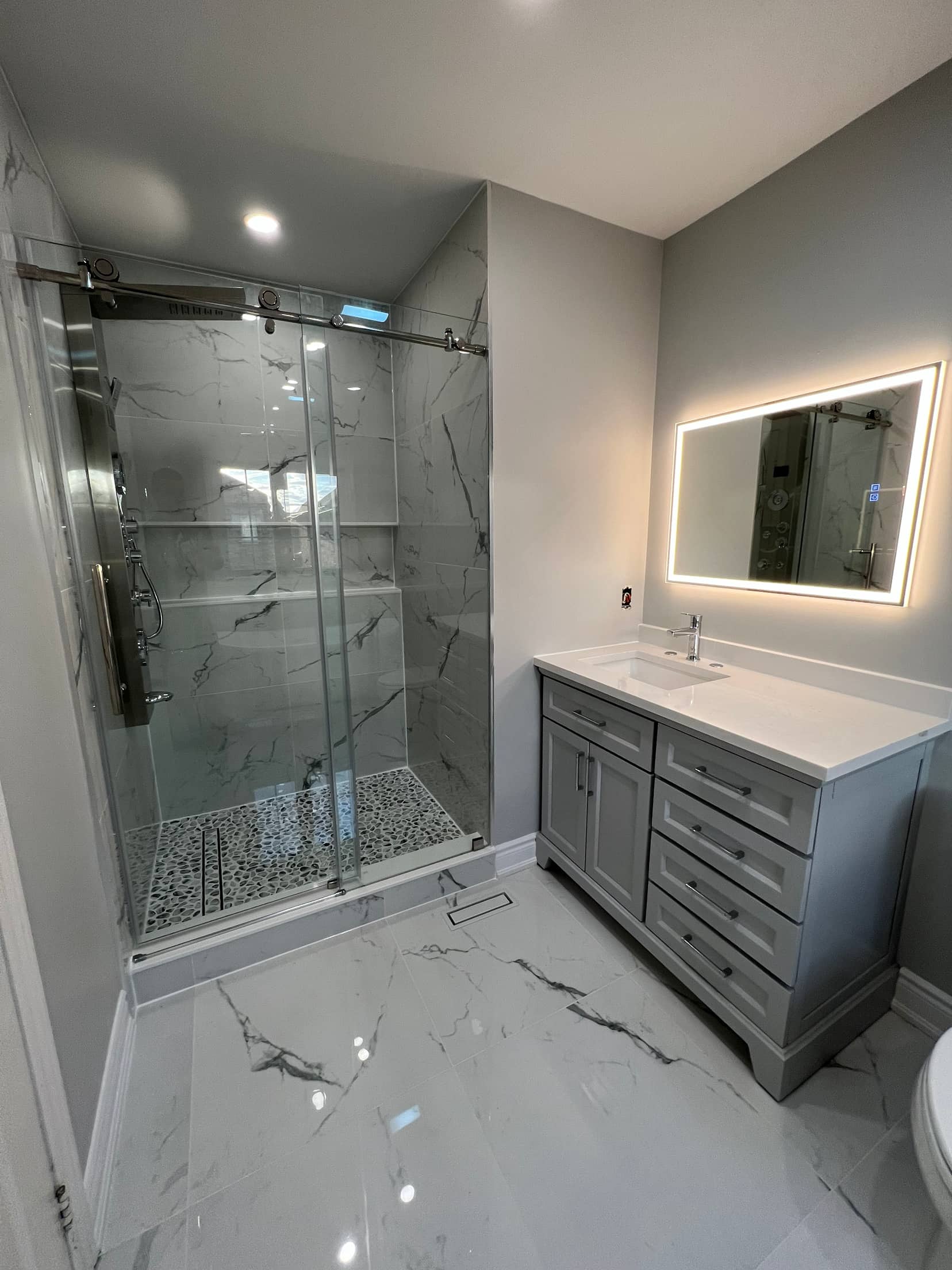 Sliding glass shower in marble look tile with a pebble mosaic floor, beside a grey vanity and backlit mirror