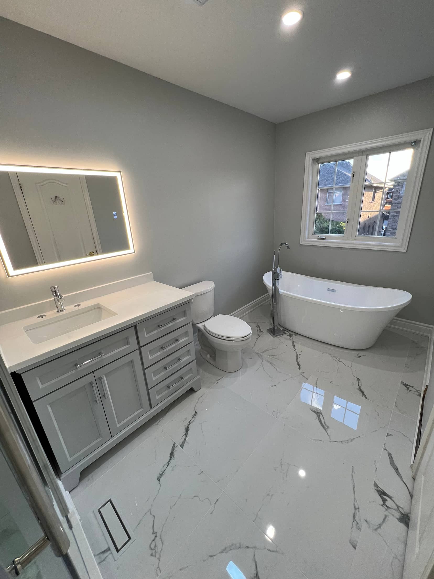 Grey shaker vanity with a backlit mirror beside a freestanding slipper tub on polished marble look tile