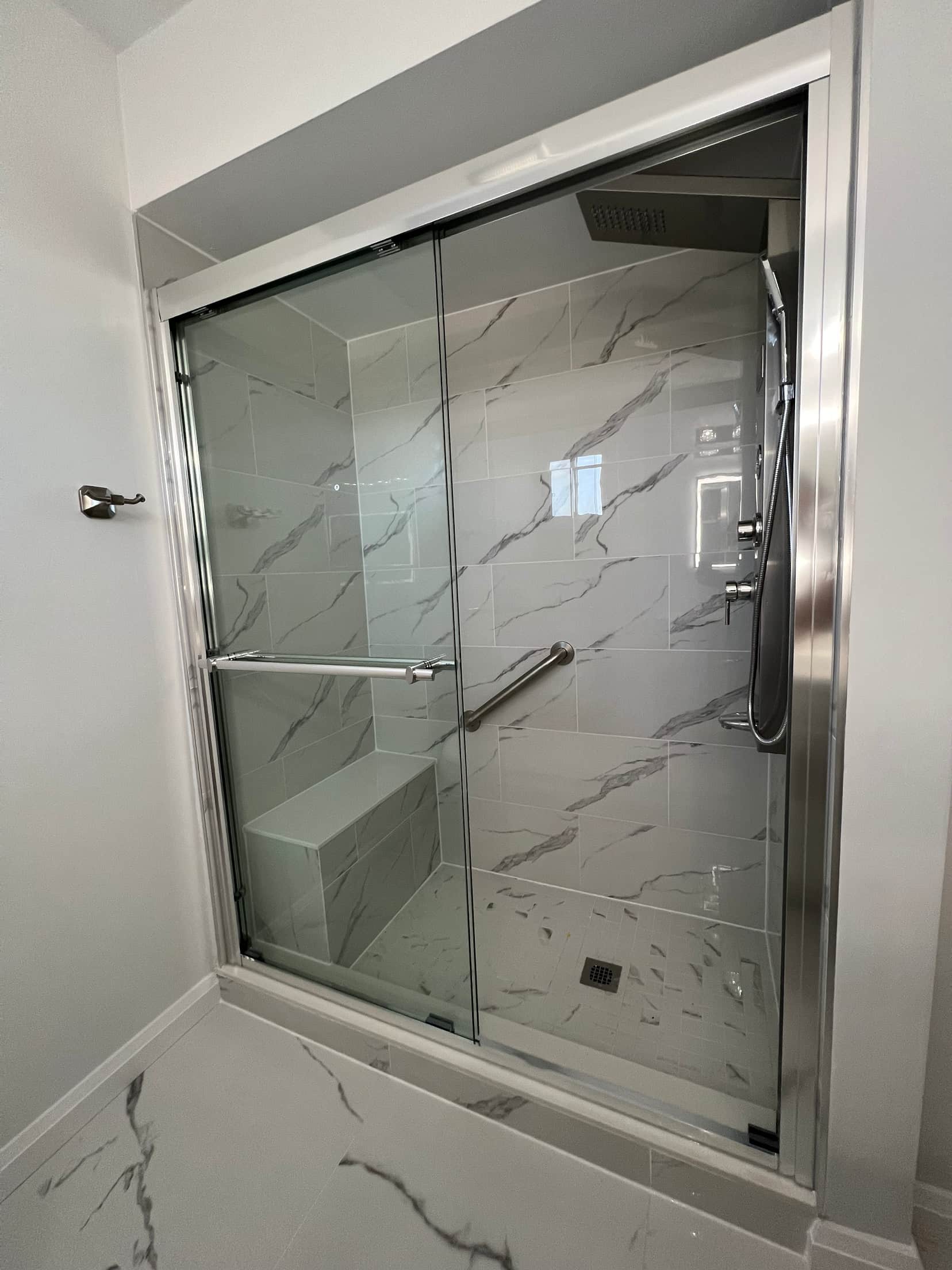 Sliding glass shower doors in a chrome frame over a marble look tiled surround with a built in bench