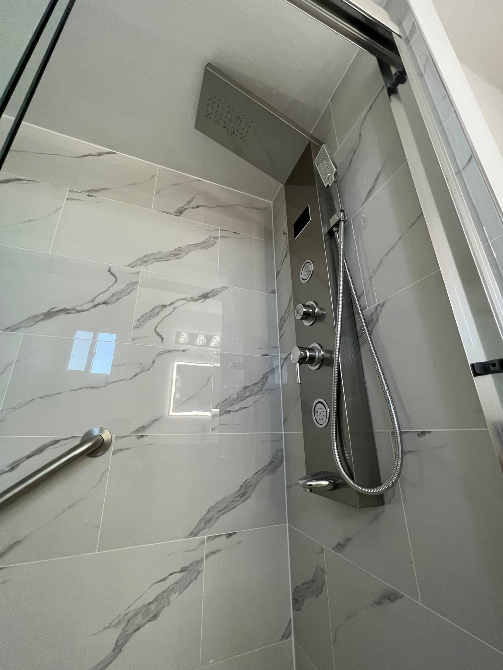 Stainless shower panel with a rainfall head, body jets and a handheld, set into a glossy marble look tiled wall beside a grab bar