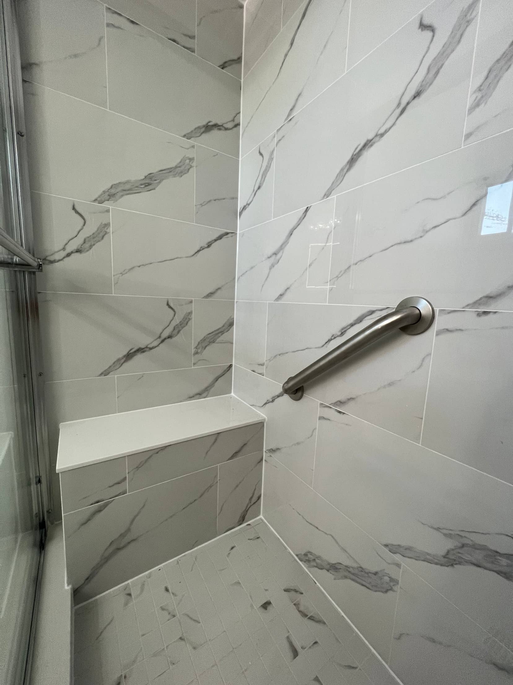 Corner of a marble look tiled shower with a white stone bench seat and a brushed nickel grab bar