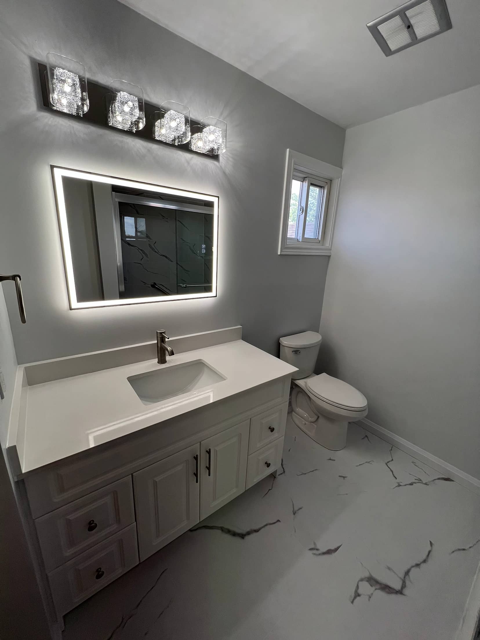 Backlit mirror under a crystal light bar above a white vanity, on marble look floor tile