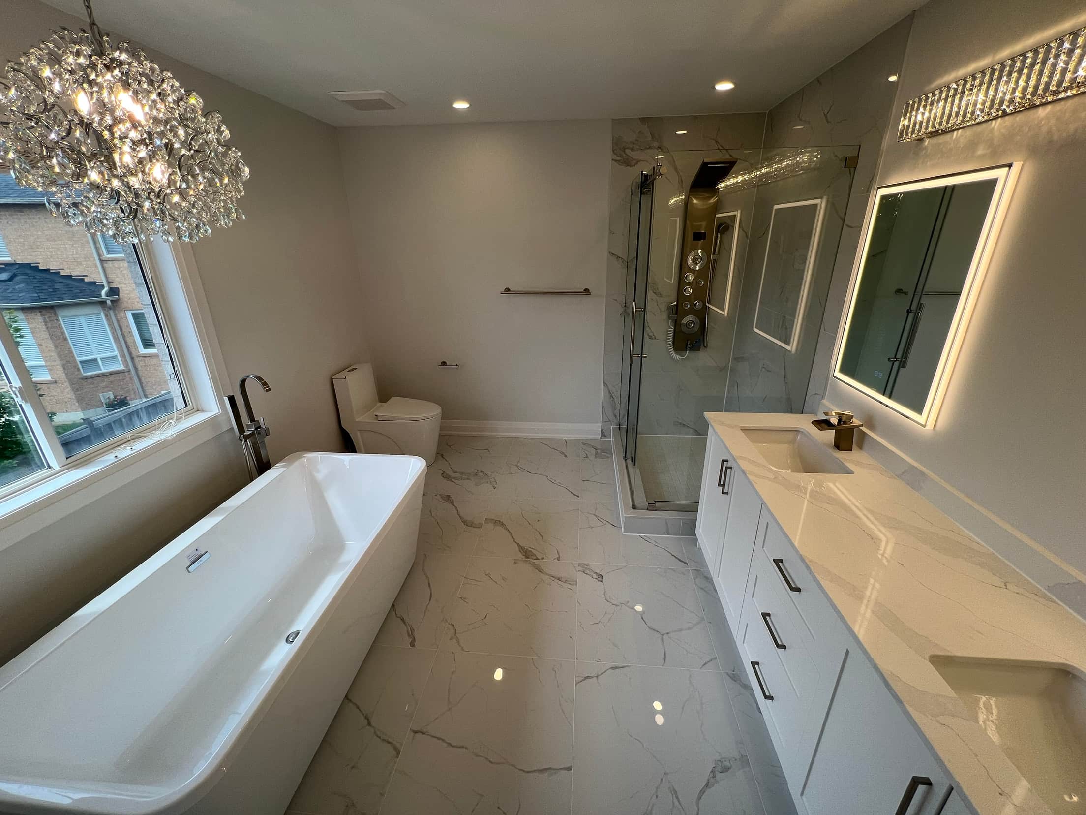 Freestanding tub beside a window under a crystal chandelier, with a glass shower and a white double vanity on polished marble floor