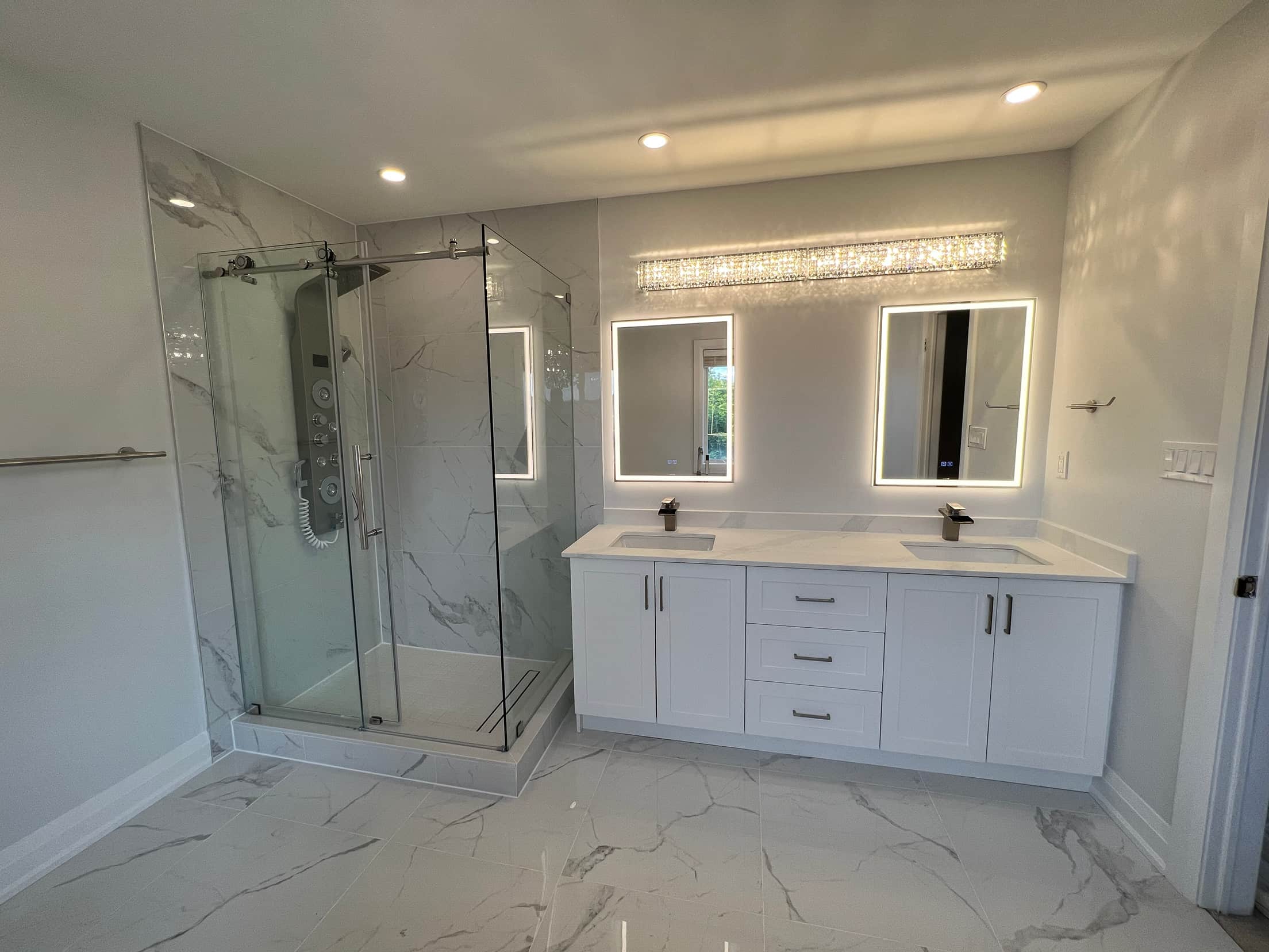 Frameless glass shower with a marble look surround and shower panel, beside a white double vanity with backlit mirrors