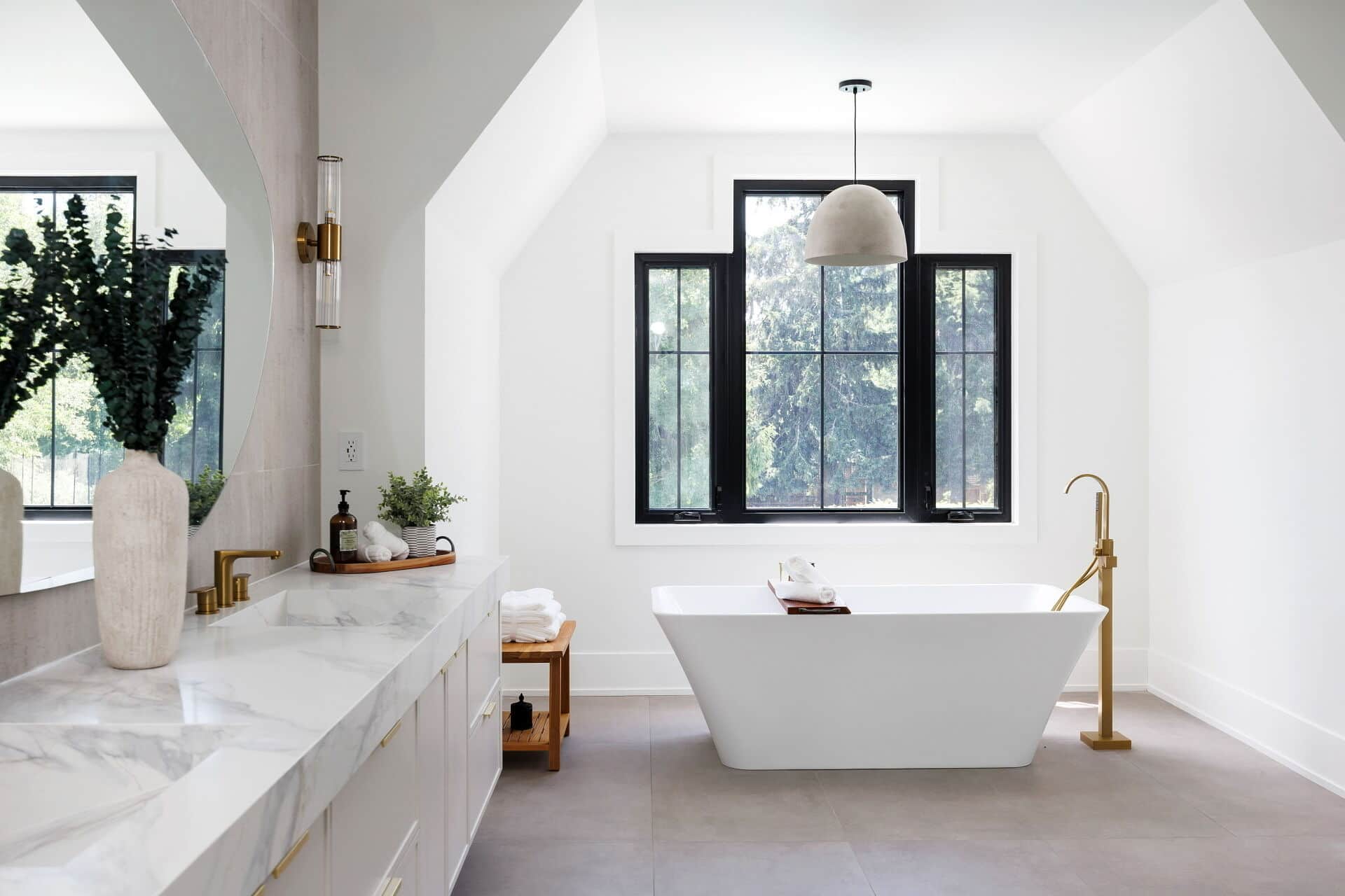 Freestanding tub beneath black framed windows, with a marble vanity run, brass filler and a teak bench