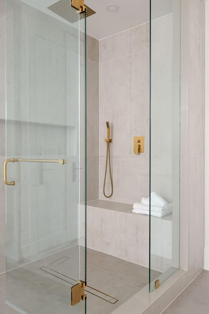 Walk in shower in pale travertine look tile with a brushed gold rainfall head, handheld and valve, a built in bench and a linear drain behind frameless glass
