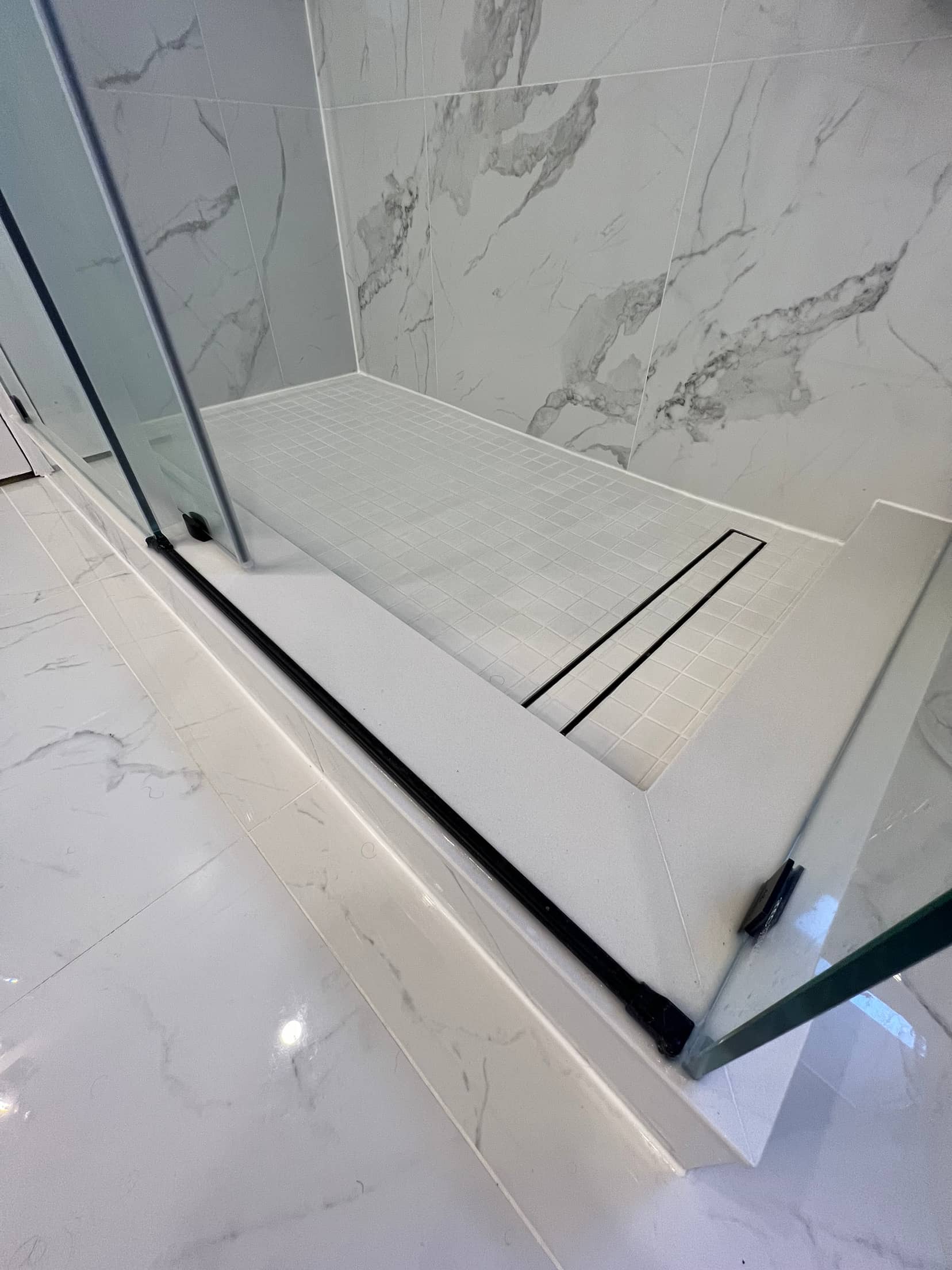 Curbless shower with a square mosaic tiled floor, a black linear drain and frameless glass against a marble look surround