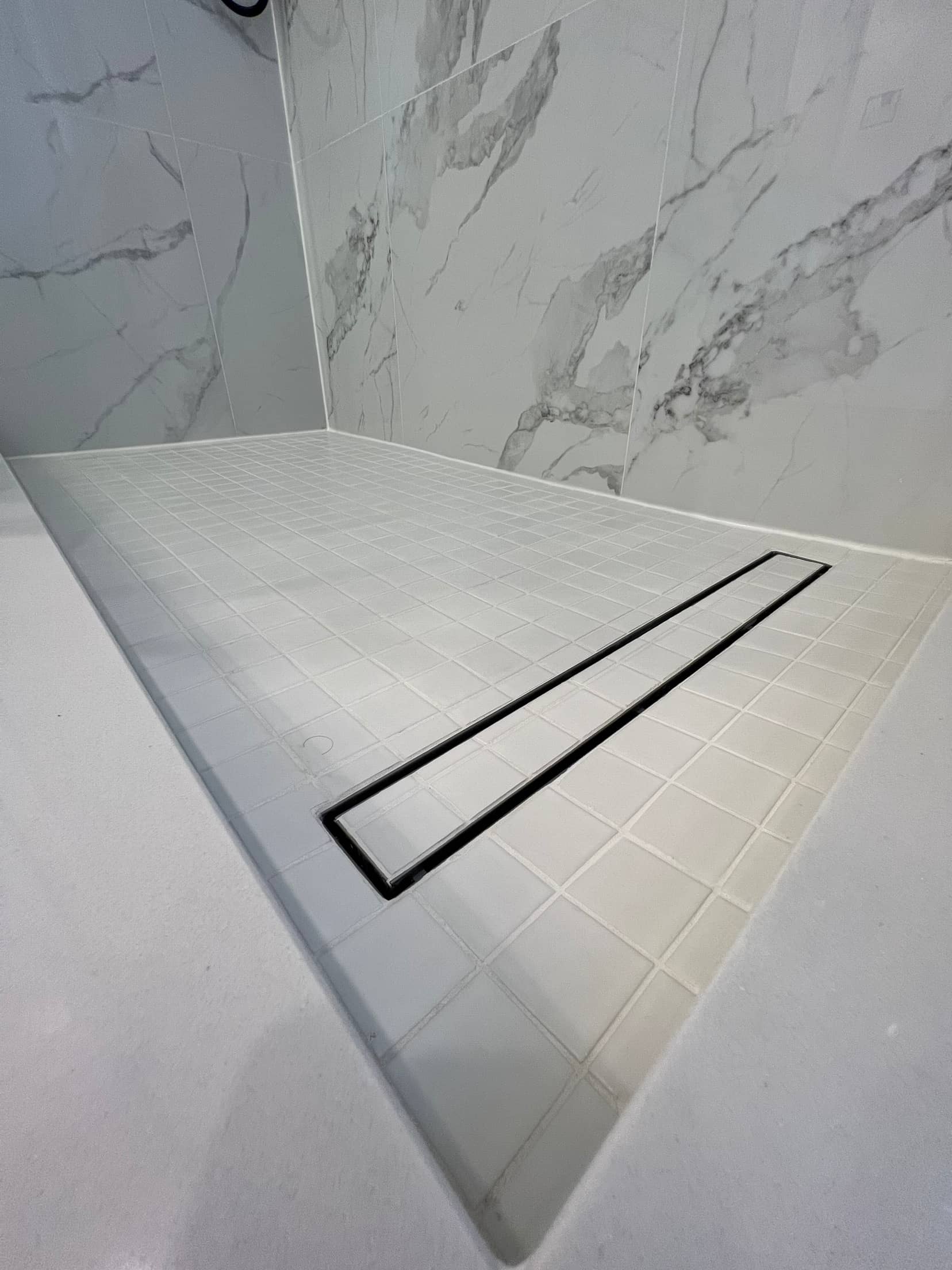 Close up of a curbless shower floor in square mosaic tile with a black linear drain, against book matched marble look walls
