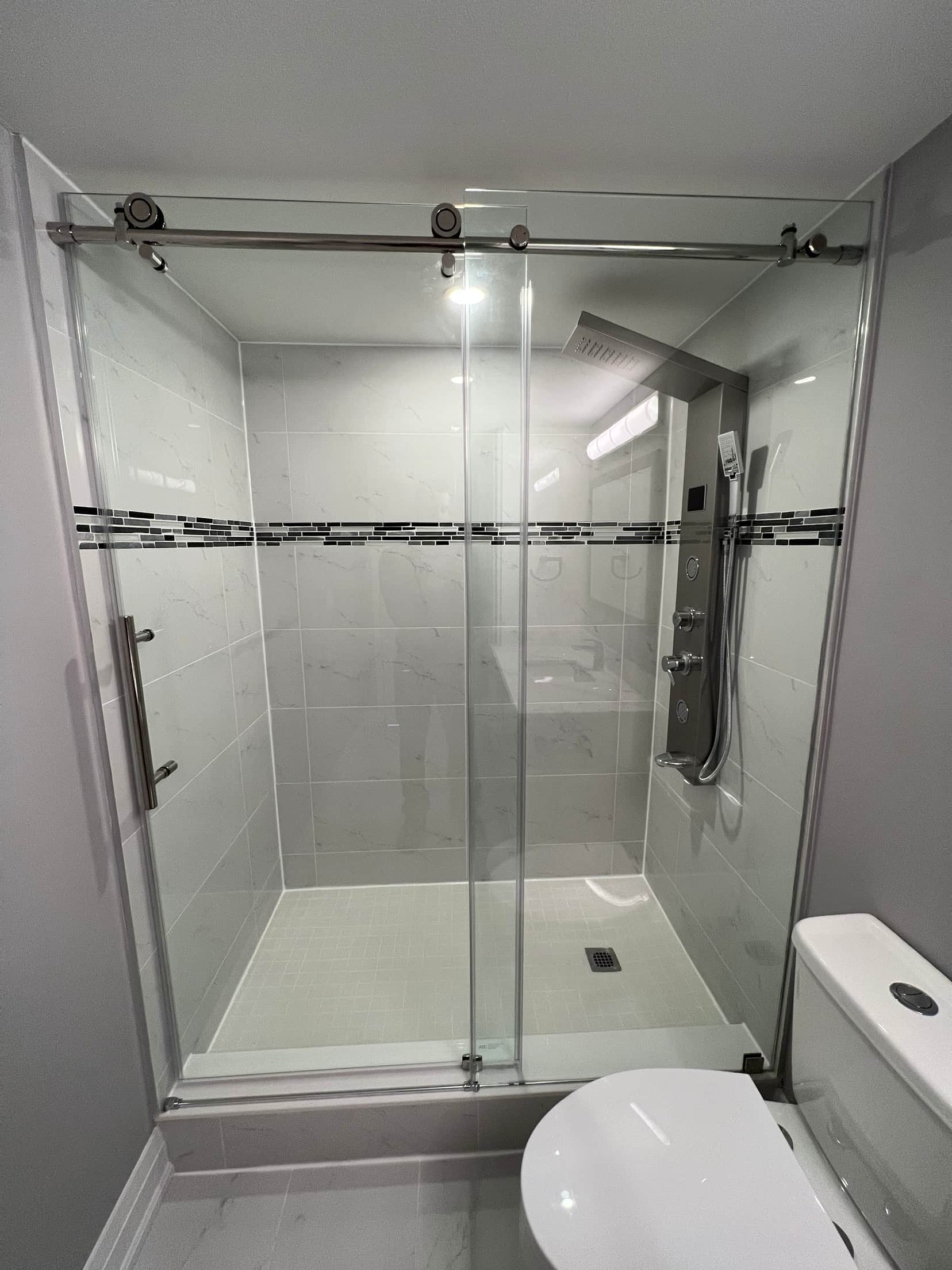 Sliding glass shower enclosure in marble look tile with a dark mosaic accent band and a stainless shower panel