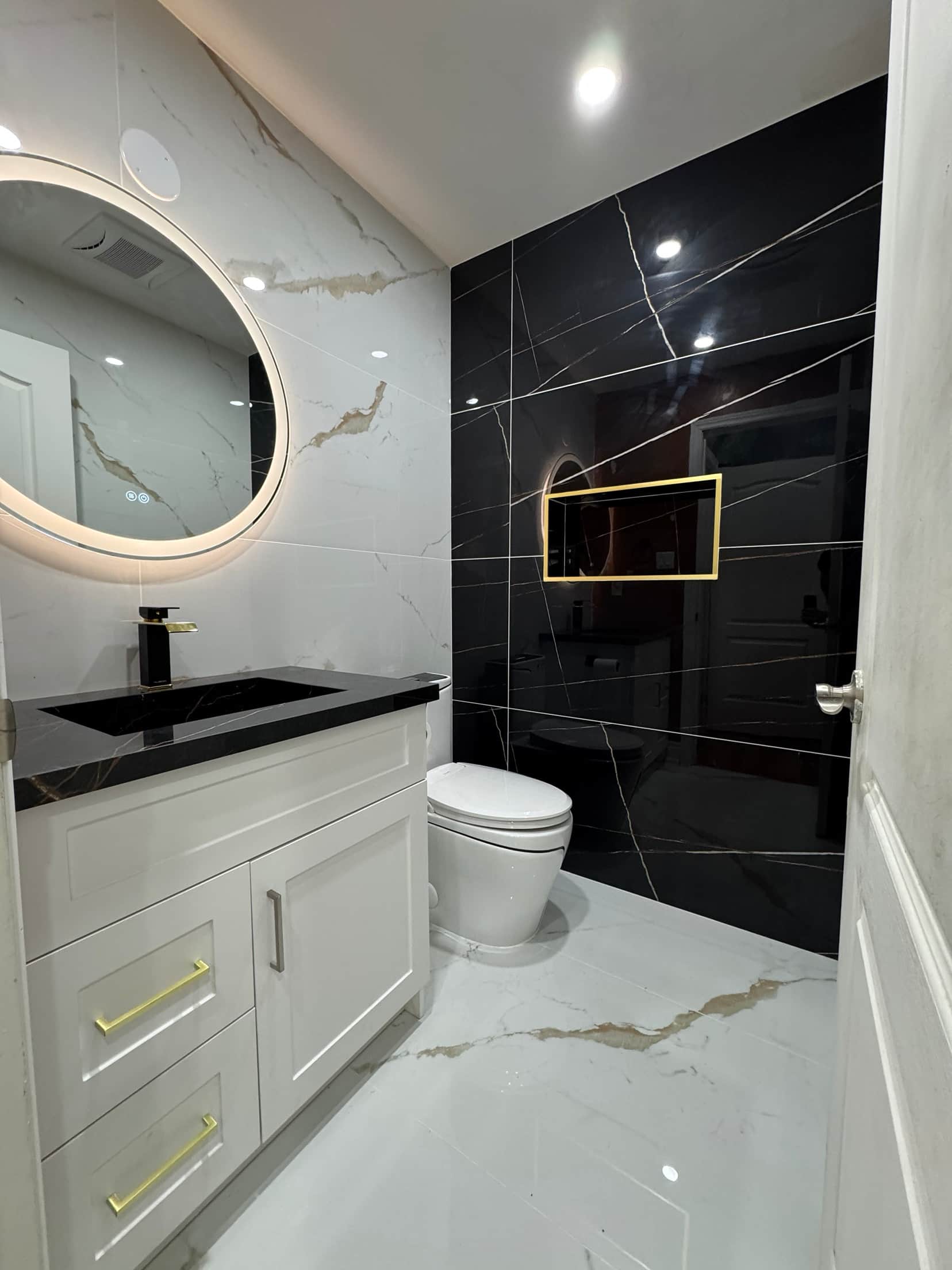 Powder room with a backlit round mirror, black stone vanity top and a black marble feature wall