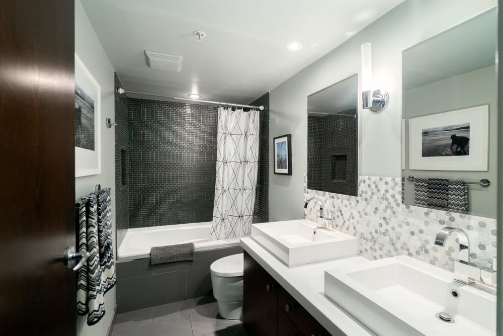 Condo bathroom with a dark mosaic tub surround, twin vessel sinks and a patterned tile backsplash