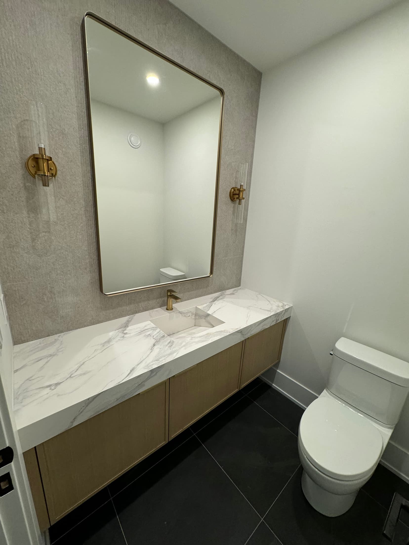 Powder room with a floating oak vanity, marble waterfall top, brass mirror and sconces over dark floor tile