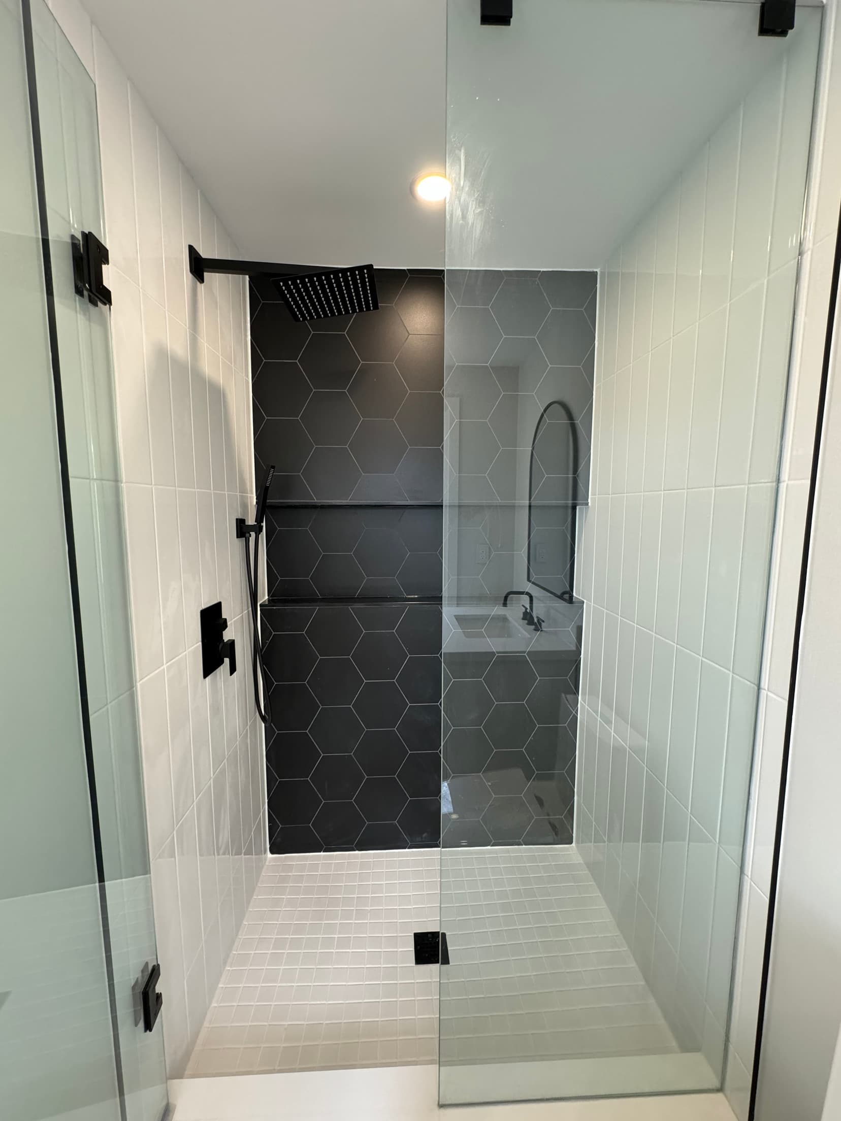 Walk in shower with a black hexagon tiled feature wall, a matte black rainfall head and handheld, white stacked tile side walls and frameless glass